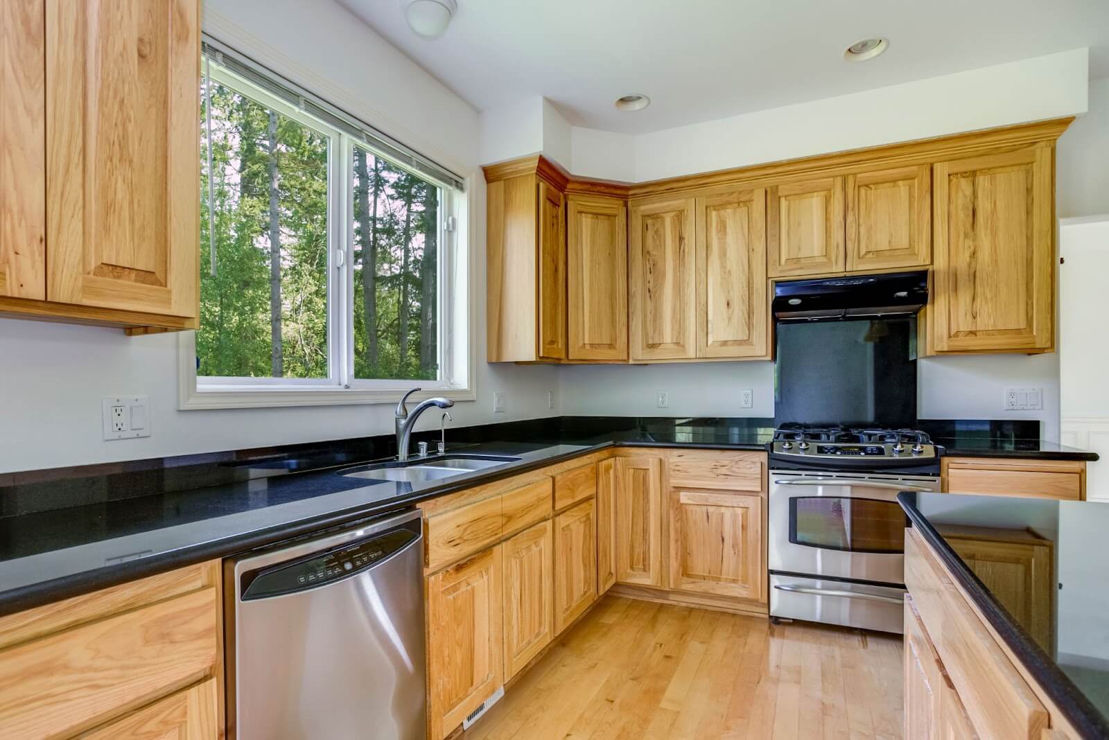 Bright kitchen with light wooden cabinets, black countertops, stainless steel appliances, and a view of trees through a large window.