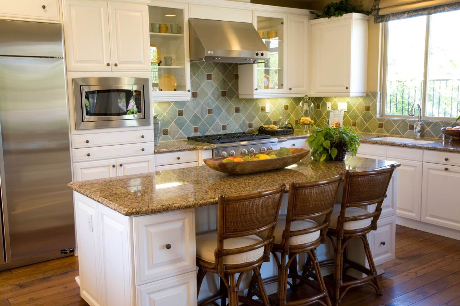 Bright kitchen with white cabinets, granite island, wicker stools, and a bowl of fruit under pendant lights.