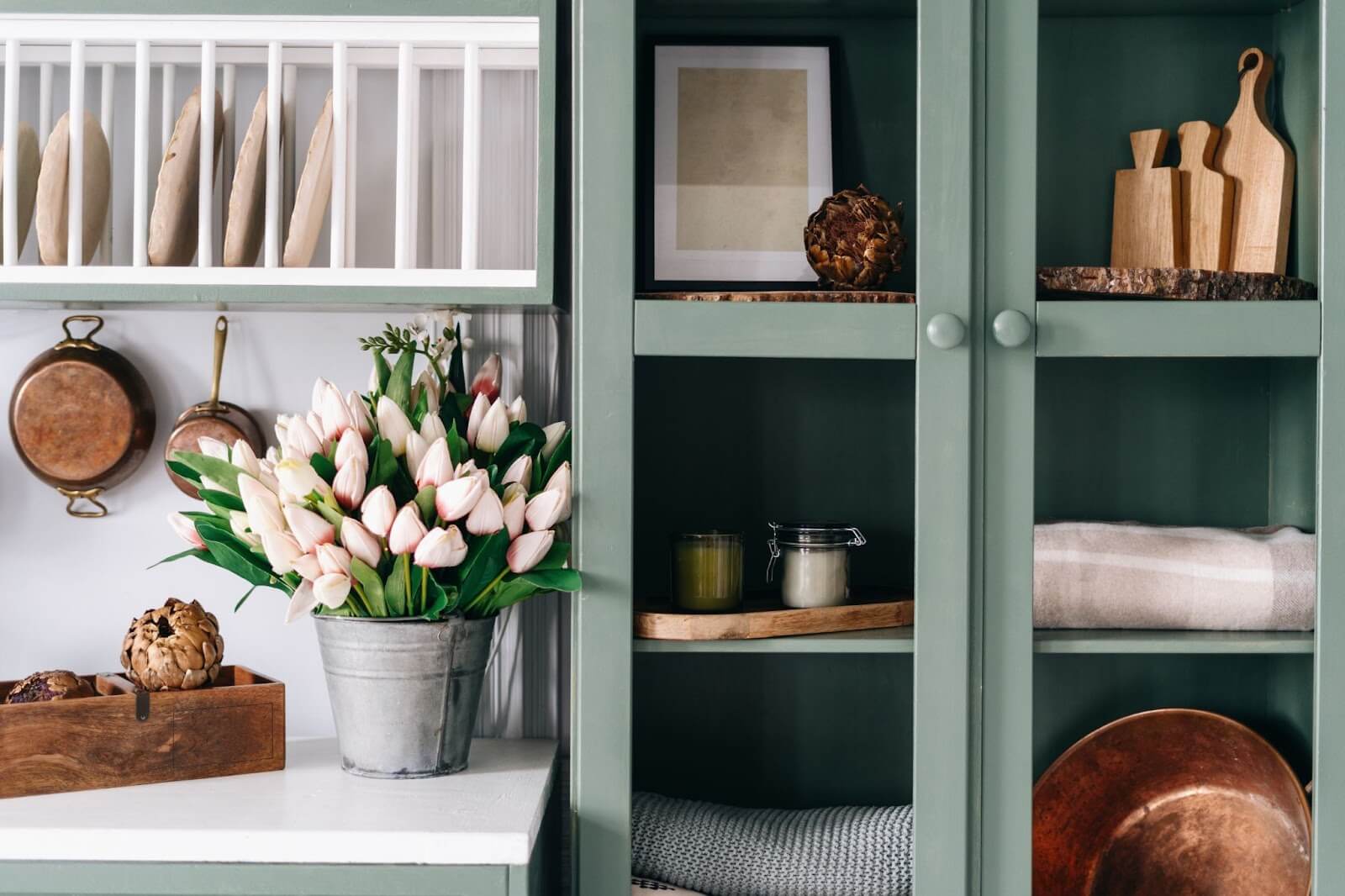 Green kitchen cabinet with glass doors, displaying flowers, candles, cutting boards, and a blanket, next to a plate rack and copper pots.