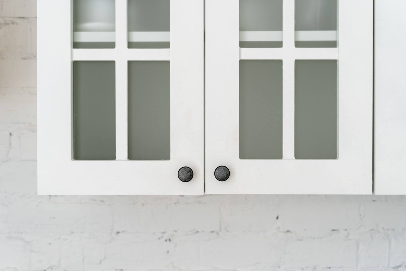 White kitchen cabinet with frosted glass doors and black knobs against a white brick wall.