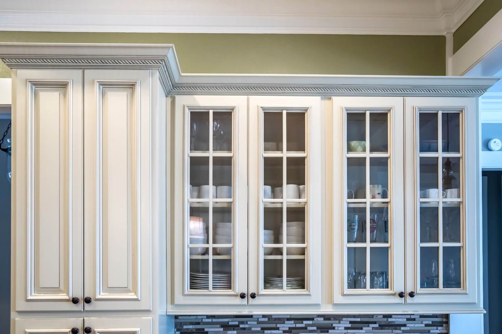 White kitchen cabinets with glass doors, displaying dishes and glassware, against a green wall with a mosaic tile backsplash.